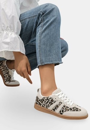 Personne portant un jean bleu et des baskets blanches à imprimé léopard, pointant vers la chaussure en s'accroupissant sur un fond gris.