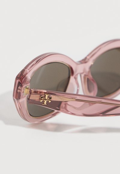 Στρογγυλά διάφανα ροζ γυαλιά ηλίου με σκούρους φακούς και χρυσή λεπτομέρεια λογότυπου στο πλαϊνό μπράτσο, τοποθετημένα πάνω σε μια λευκή επιφάνεια.