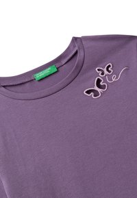 Fialové tričko s kulatým výstřihem a dvěma vyšitými motivy motýlů poblíž výstřihu, se zeleným štítkem "United Colors of Benetton" uvnitř límce.