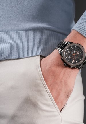 Miehen käsi, jossa musta metallinen rannekello näkyvine viisareineen, käsi beigen väristen housujen taskussa ja näkyvissä vaaleansininen ribbineulostakki.