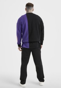 Felpa nera e viola con design a spacco, polsini a costine e una maglietta nera a strati. Abbinata a pantaloni neri e sneaker bianche.