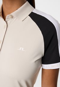 Ljusbeige polo-shirt med en texturerad krage, som har svart och vita detaljer på ärmarna och en liten vit logotyp på bröstet.