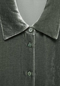 Coleto de camisa de terciopelo verde oscuro con cinco botones. Textura suave, brillo prominente y un acabado de tela suave. Diseño clásico con una apariencia pulida.