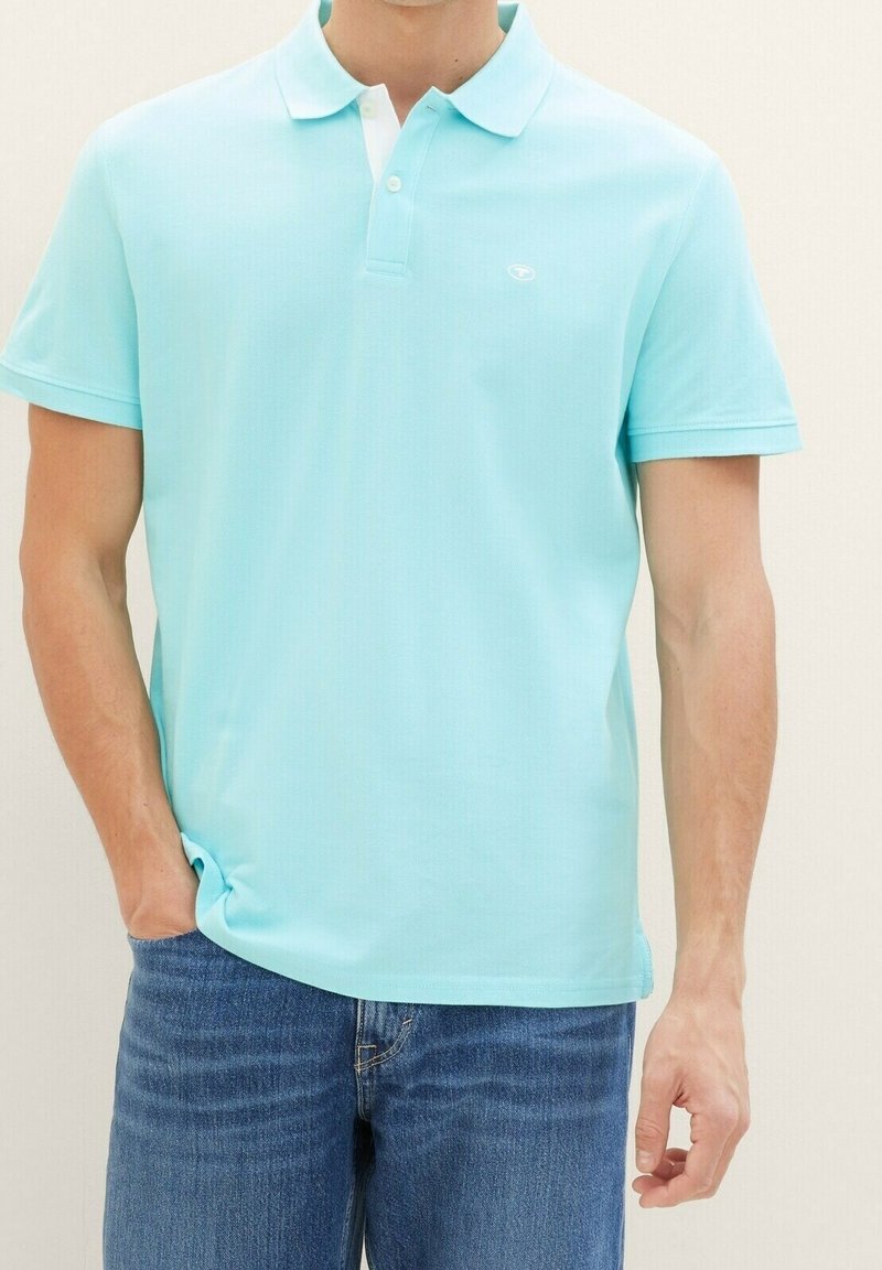 Polo shirt bleu clair avec une finition texturée, manches courtes et col blanc. Présente un petit logo sur la poitrine. Porté avec un jean en denim.