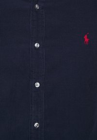 Granatowa koszula z sztruksu z sześcioma srebrnymi guzikami i małym czerwonym haftowanym logo po lewej stronie. Tkanina o fakturze w pionowe paski.