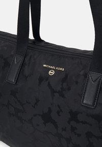 Musta Michael Kors -kassi, jossa on hillitty kukkakuosi, kullanvärinen logo-plaatti, vetoketjuku closure ja kaksi kangaskahvaa.