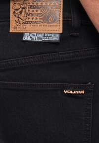 Sort denim med et lædermærke, der har et design og logo, samt et etikette, der angiver vandbesparende foranstaltninger. Subtile stikningsdetaljer.