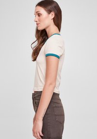 Short-sleeve beige t-shirt with turquoise trim, featuring a fitted design and soft texture. Paired with high-waisted brown jeans.