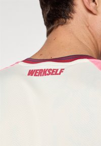 Maillot de sport blanc avec un design texturé, accentué par un col rouge et bleu. Le dos affiche "WERKSELF" en lettres rouges en gras.