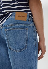 Ljusblå denimjeans med rak bendesign, med en tanfärgad läderetikett i midjan och två bakfickor. Randig skjorta synlig ovanför.