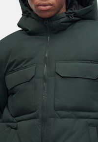 Gros plan d’une personne portant une veste matelassée à capuche vert foncé avec une fermeture éclair à l’avant et deux poches à rabat sur la poitrine.