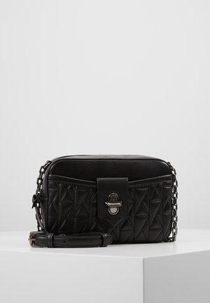 Sac bandoulière en cuir noir matelassé avec un rabat avant et un fermoir tourniquet argenté, ainsi qu'une bandoulière chaîne réglable, posé sur une surface blanche.