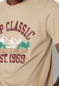 Beżowa bawełniana koszulka z grafiką przedstawiającą góry, drzewa oraz tekst "CAMP CLASSIC EST. 1969" w kolorze bordowym.