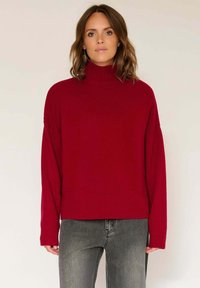 Pull en tricot rouge avec un col montant, des épaules tombantes et un ourlet côtelé. Associé à un jean gris, présentant une silhouette décontractée et un contraste de textures.