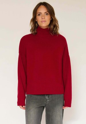 Pull en tricot rouge avec un col montant, des épaules tombantes et un ourlet côtelé. Associé à un jean gris, présentant une silhouette décontractée et un contraste de textures.