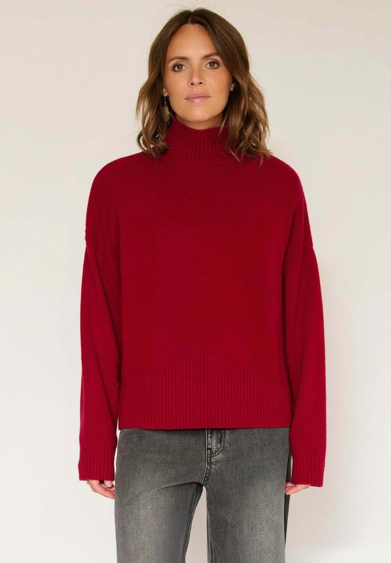Pull en tricot rouge avec un col montant, des épaules tombantes et un ourlet côtelé. Associé à un jean gris, présentant une silhouette décontractée et un contraste de textures.