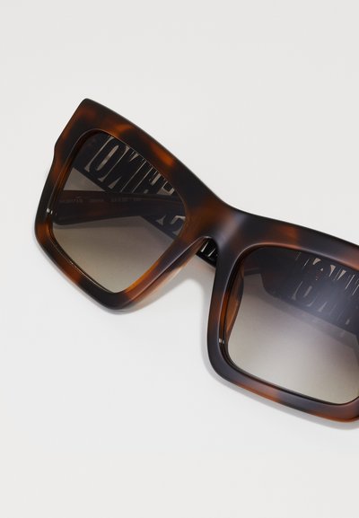 Ochelari de soare supradimensionați maro cu model țestoasă, dotati cu lentile cu gradient și o ramă groasă, lucioasă. Interiorul prezintă detalii de design cu marcă.