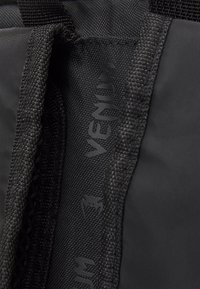 Črna športna torba z teksturirano tkanino, mrežnimi detajli in natisnjenim logotipom "VENUM" v črni barvi. Trajen material z ojačenimi šivi.