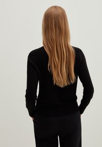 Černý pletený svetr s dlouhými rukávy, přiléhavý střih a žebrovaný lem, kombinovaný s tmavými kalhotami. Pohled zezadu odhaluje dlouhé rovné vlasy.