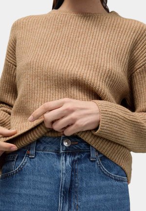 Persona con un suéter de punto beige acanalado y vaqueros azules, ajustando el borde inferior del suéter con ambas manos.