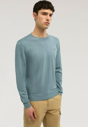 Joven con suéter azul claro de cuello redondo y pantalones cargo beige, de pie con una mano en el bolsillo frente a un fondo liso.