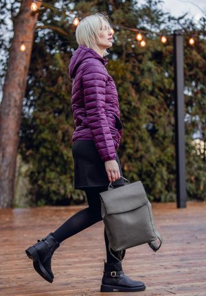 Nafoukaná fialová bunda, černá sukně a černé kozačky nad kolena. Šedý batoh s klopou a ramenními popruhy. Pozadí z dřevěné terasy.