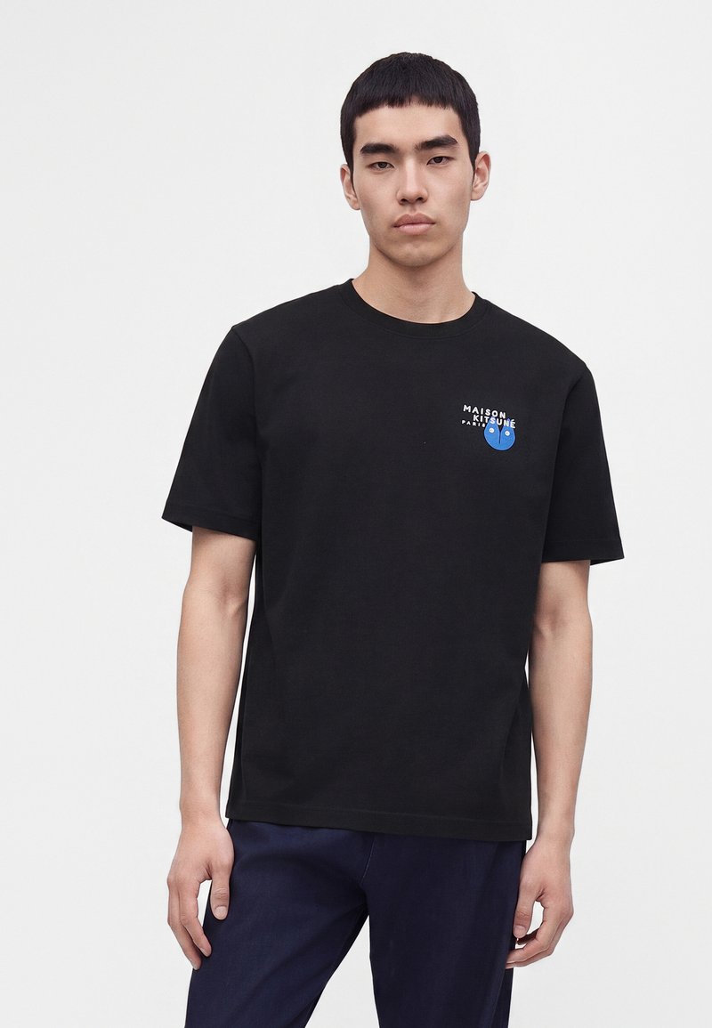 Νέος άνδρας που φοράει μαύρο T-shirt Maison Kitsuné με μικρό μπλε λογότυπο αλεπούς στο στήθος, στέκεται μπροστά σε ένα απλό φωτεινό φόντο.