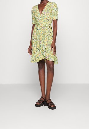 Femme portant une robe portefeuille jaune à manches courtes, col en V, imprimé floral multicolore, et des sandales compensées noires.