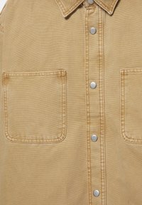 Chemise beige à boutons avec deux poches carrées sur le devant et des boutons-pression argentés le long de la patte centrale.