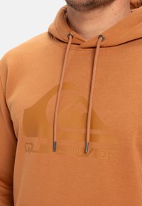 Pullover hoodie i lys orange stof med et gummilogo i midten. Justerbare snore med metalspidser, blød tekstur.
