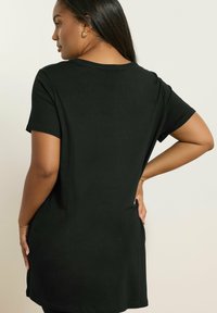 Nainen, jolla on pitkät tummat hiukset, pukeutuneena yksinkertaiseen mustaan lyhythihaiseen tunikamalliseen paitaansa, näkyy selkäpuolelta seisten käsi lanteella.