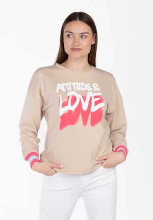 Kobieta w beżowej bluzie z napisem „ATTITUDE & LOVE” w białych i różowych literach, w białych spodniach, z rękami w kieszeniach, patrząca w prawo.