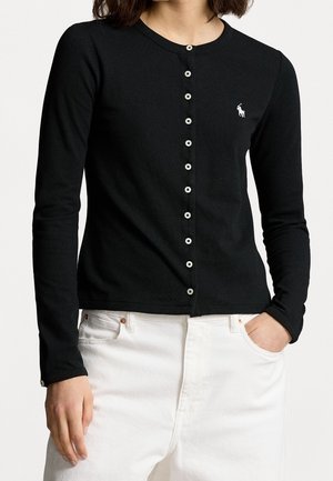 Kvinde iført en sort knap-lukket cardigan med lange ærmer og en lille hvid logo på brystet samt hvide bukser, med den ene hånd i lommen.