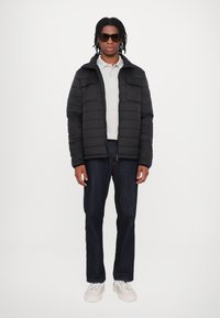 Giacca padded nera con chiusura a zip, due tasche frontali, indossata sopra un polo grigio chiaro. Abbinata a jeans scuri e sneakers bianche.