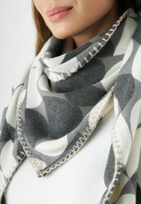 Femme portant un foulard à motifs gris et blanc avec des bords cousus sur un haut blanc, ne montrant que la partie inférieure du visage et le cou.