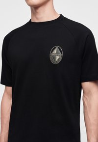 Άνδρας που φοράει μαύρο T-shirt με μεταλλικό έμβλημα που φέρει τη λέξη "HUGO" στην αριστερή πλευρά του στήθους.