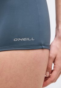 Κοντινό πλάνο μπλε μαγιό O'Neill, φορεμένο στο πάνω μέρος του μηρού δίπλα σε ένα χαλαρό χέρι.