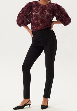 Kobieta ubrana w przezroczystą bordową bluzkę z kwiatową koronką i bufkami, dopasowane czarne spodnie o wysokim stanie oraz czarne czółenka na obcasie typu kitten heel z paskiem z tyłu.