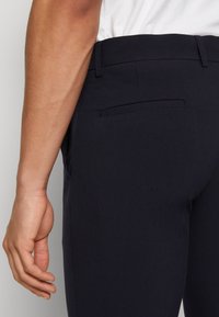 Marinblå byxor med en slät yta, slim fit, bältesloopar och detaljer i bakfickan. Tyget verkar vara lätt och strukturerat.