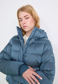 Nafučana plava jakna s kapuljačom, koja sadrži dugmad za zatvaranje i rebraste manžete, izrađena od sjajnog materijala s kviltanim detaljima.