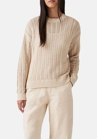 Donna che indossa un maglione beige a trama aperta con maniche lunghe e polsini a coste, abbinato a pantaloni beige chiaro, in piedi contro uno sfondo bianco.