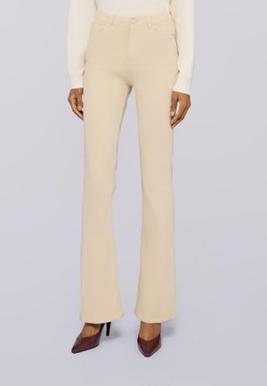 Slanke beige hoge taillebroeken gedragen met een witte lange mouwen top en donkergroene spitse hoge hakken op een lichtgrijze achtergrond.