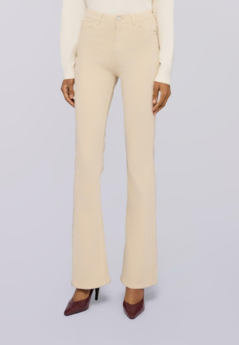 Pantalones beige de talle alto y ajustados combinados con una blusa blanca de manga larga y zapatos de tacón alto puntiagudos marrón oscuro sobre un fondo gris claro.