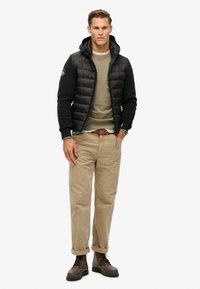 Sort puffer vest med hætte over en olivengrøn sweater, beige chinos og brune snørebåndsstøvler. Har rullede ærmer og et ur.
