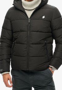 Musta toppatakki, jossa vetoketju edessä, huppu ja sivutaskut. Takissa on tikattuja osioita ja valkoinen salaman muotoinen logo rinnassa.