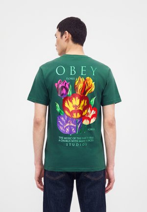 Hombre con camiseta verde con flores de tulipán coloridas y texto "OBEY" impreso en la espalda, combinado con vaqueros azul oscuro.