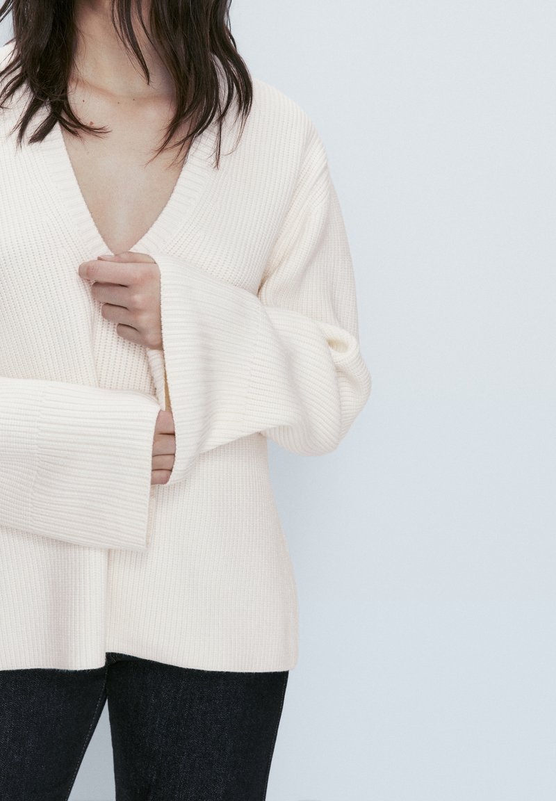 Pull en maille côtelée crème avec un col en V profond, des manches larges et un tissu texturé. Associé à un jean foncé.