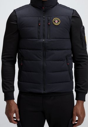 Mand iført sort langærmet skjorte og sort quiltet lynlåsvest med gul cirkulær Zavetti Canada-logo på brystet.