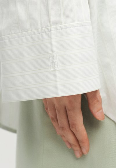 Main reposant sur le côté, portant une chemise blanche à manches longues avec de fines rayures horizontales et un poignet monogrammé, associée à un pantalon vert clair.
