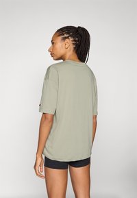 Lys grønn bomull T-skjorte med korte ermer, avslappet passform, med en logo på venstre erme, brukt med svarte shorts. Bakfra.